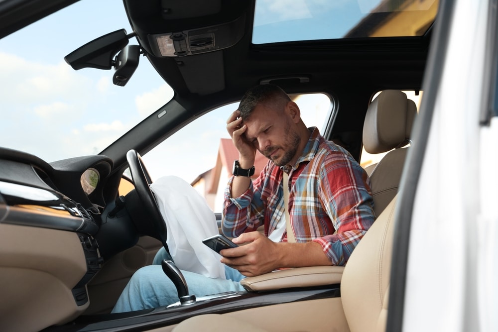 Hombre preocupado sentado en el asiento del conductor de un automóvil, mirando su teléfono móvil, reflejando el estrés y la confusión tras un accidente automovilístico.